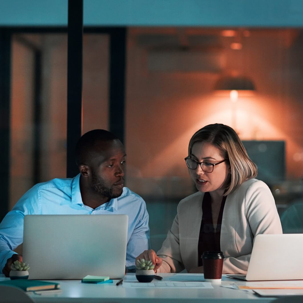 Two individuals collaborating on laptops in a modern office setting, focused on their tasks and sharing ideas.