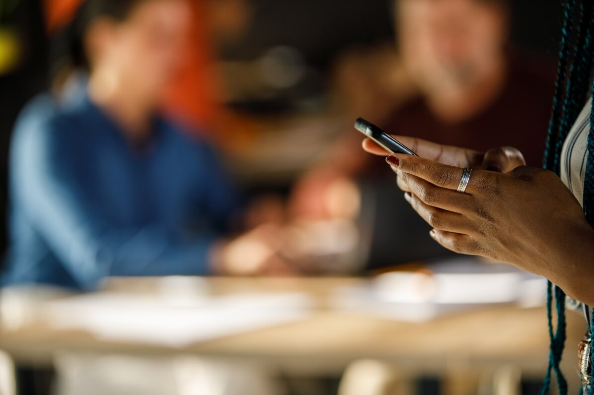 A woman seated at a table, holding a cell phone, engaged in conversation or browsing content.