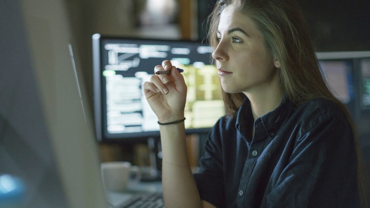 A woman seated at a desk, using a computer monitor, while holding pen.